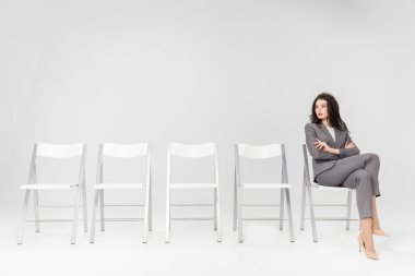 confident woman sitting with crossed arms and crossed legs on chair isolated on grey clipart