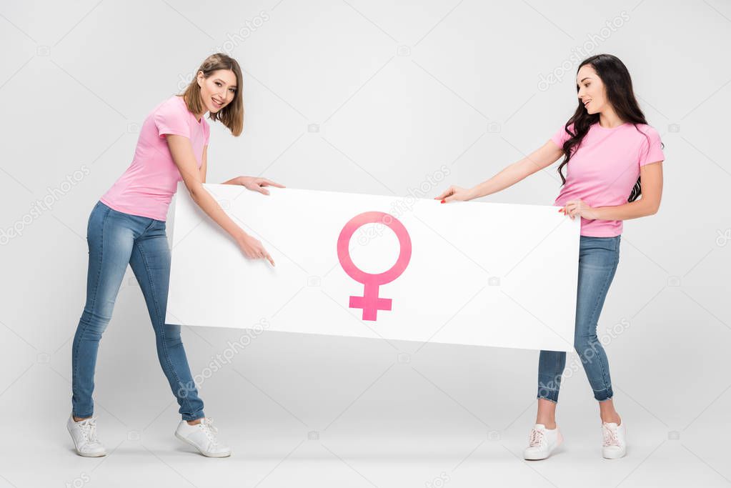 cheerful woman pointing with finger at large sign with female symbol near girl on grey background