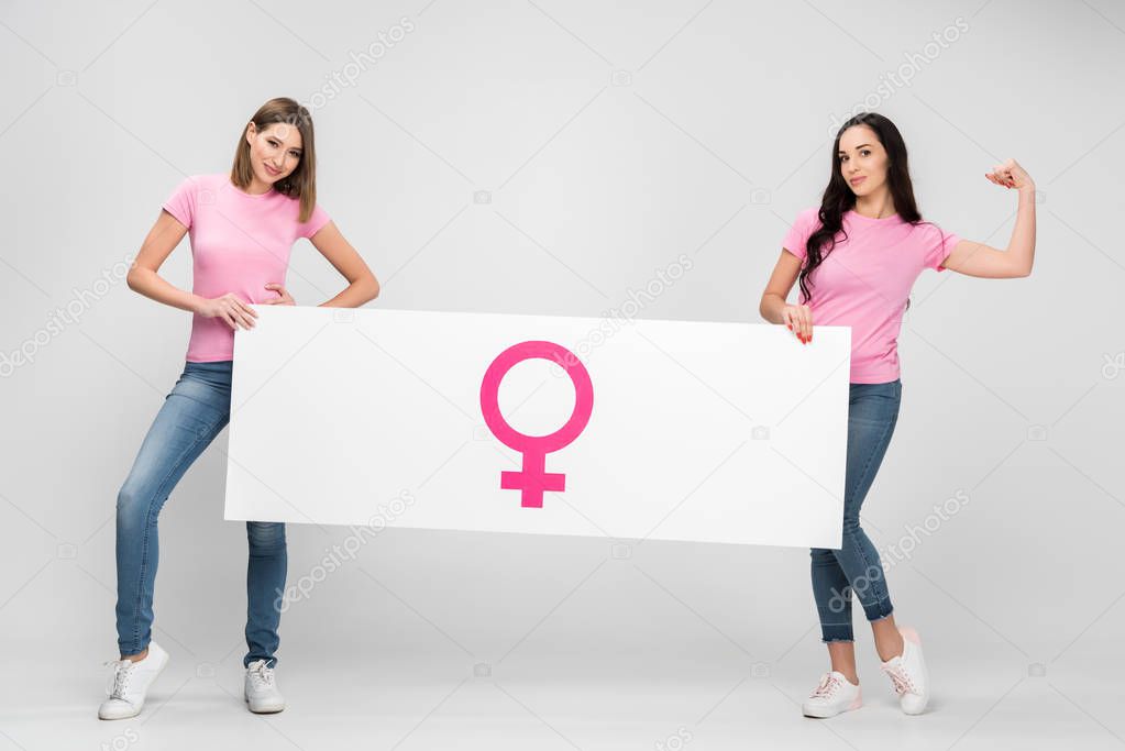 beautiful woman showing muscle and holding large sign with female symbol with friend on grey background