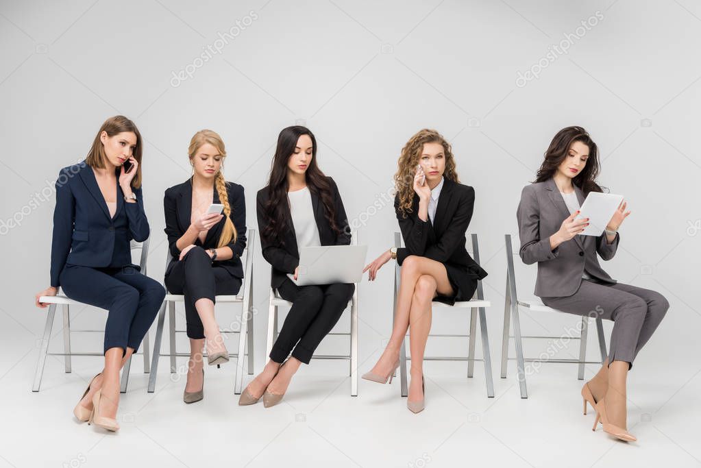 attractive colleagues in formal wear sitting on chairs and using gadgets isolated on grey
