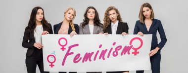 attractive businesswomen holding  large sign with feminism lettering isolated on grey  clipart