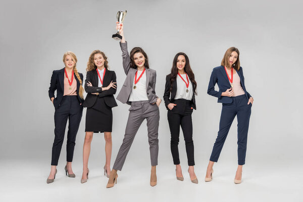 cheerful businesswoman holding trophy above head and standing near attractive colleagues posing on grey background