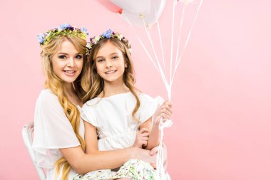 Anneler dizlerinin üstüne oturan ve pembe bir arka plan üzerinde beyaz ve pembe hava balonları tutan şirin çocuk