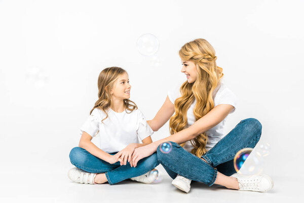 smiling mom and daughter sitting on floor with crossed legs and looking at each other while soap bubbles flying around on white background