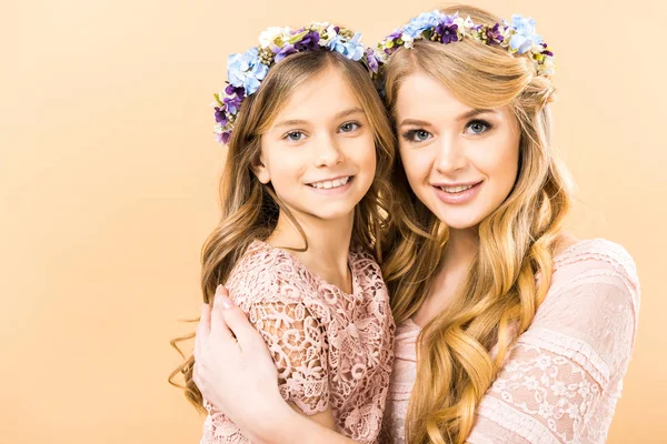 cute child and pretty mom in colorful floral wreaths embracing and looking at camera on yellow background