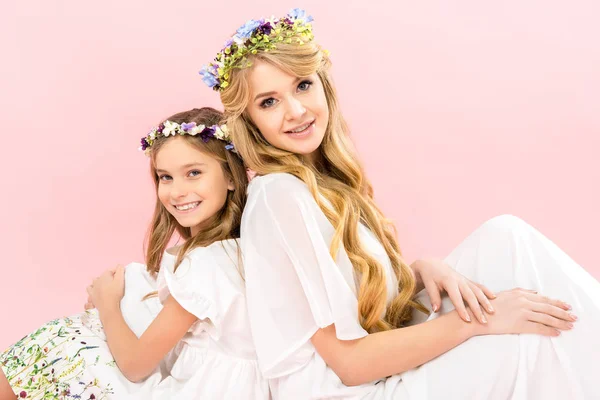 美丽的母亲和可爱的女儿在优雅的白色礼服和花卉花圈背靠背坐着 看着粉红色的背景相机 — 图库照片