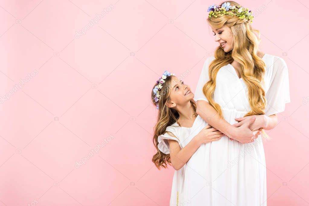 beautiful mother and adorable daughter in elegant white dresses and colorful floral wreaths on pink background with copy space