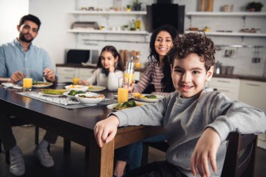 selective focus of cheerful latin son looking at camera with hispanic family on background clipart
