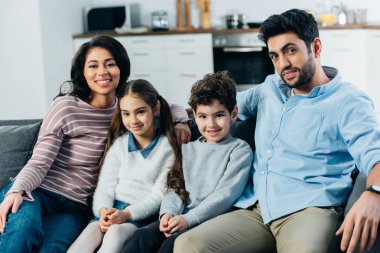 happy hispanic family sitting on sofa and smiling at home clipart