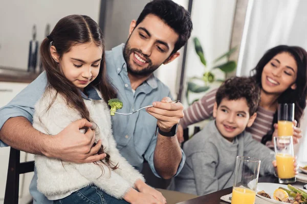 cheerful latin father feeding daughter with broccoli near wife and son at home
