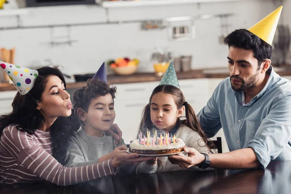 happy hispanic family in party caps blowing at candles on birthday cake at home