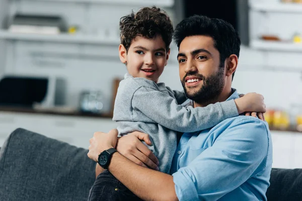 cheerful latin father hugging cute son at home