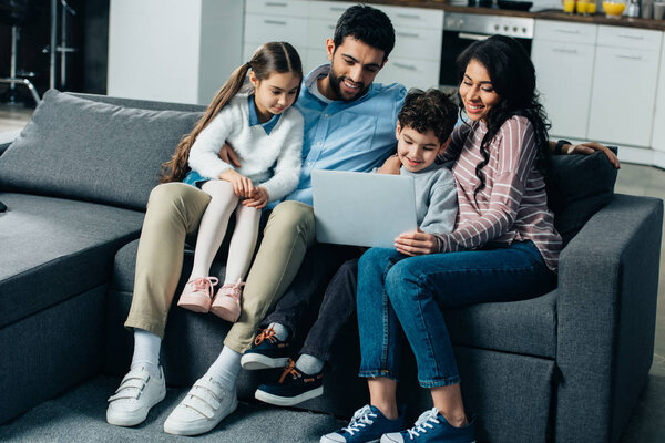 happy hispanic family sitting on sofa and looking at laptop at home