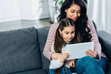 latin kadın kızıyla kanepede oturan ve evde dijital tablet kullanma
