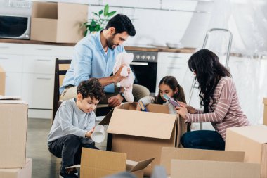 happy latin father holding soft toy near hispanic family while unpacking boxes in new home  clipart