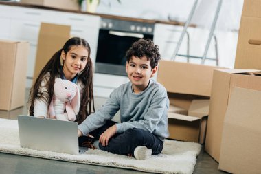 happy kids using laptop while sitting on carpet in new home clipart