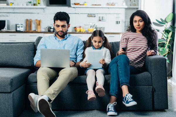 latin woman sitting on sofa near husband with laptop and daughter with digital tablet 