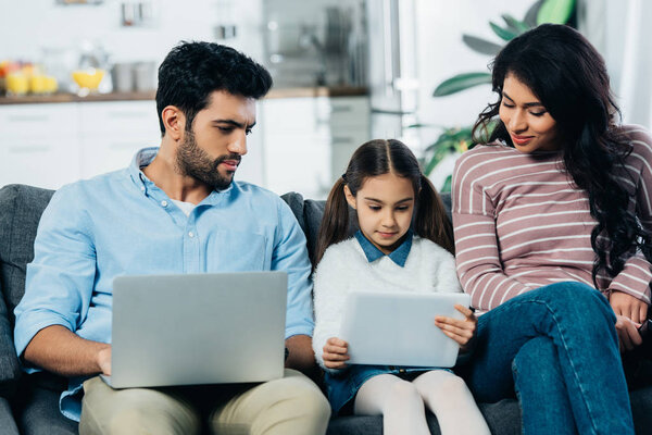 handsome latin man with laptop looking at digital tablet in hands of cute daughter sitting near mother on sofa