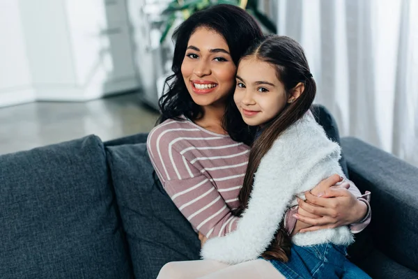 latin woman hugging cute daughter while sitting on sofa at home