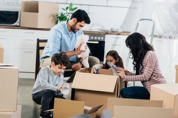 happy latin father holding soft toy near hispanic family while unpacking boxes in new home 
