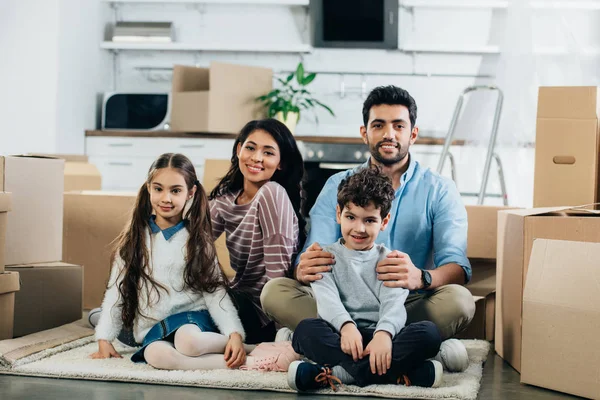 幸せなラテン夫と新しい家のかわいい子どもたちとカーペットに座って妻 — ストック写真