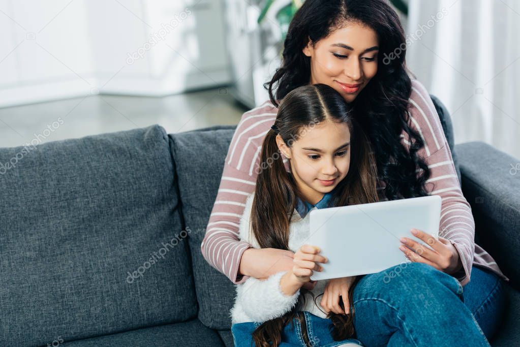 latin woman sitting on sofa with daughter and using digital tablet at home