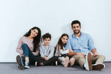 cheerful hispanic family smiling while sitting on floor near white wall  clipart