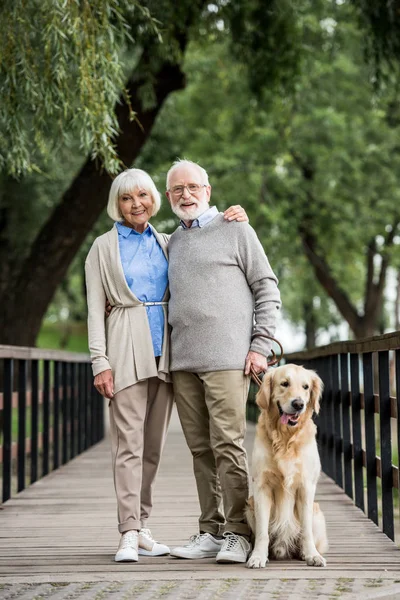 smiling senior couple hugging while walking with dog across wooden bridge