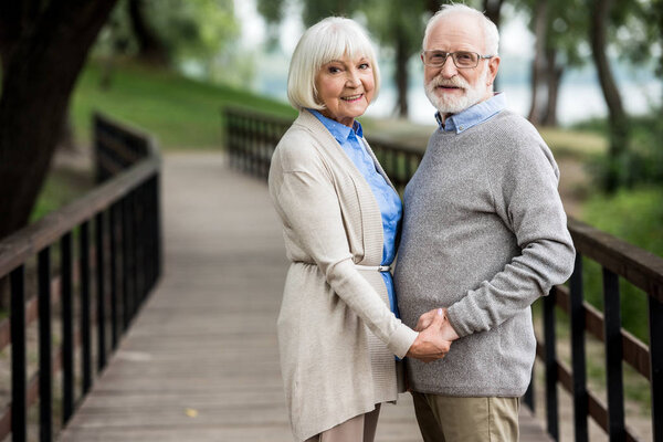 счастливая пожилая пара, стоящая на деревянном мосту и держась за руки
 