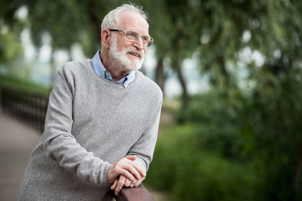 selective focus of smiling senior man in grey pullover and glasses standing by wooden bridge railing and looking away 