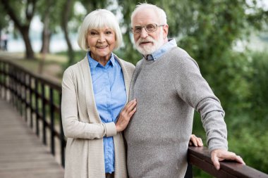 selective focus of nice smiling senior couple standing on wooden bridge in park