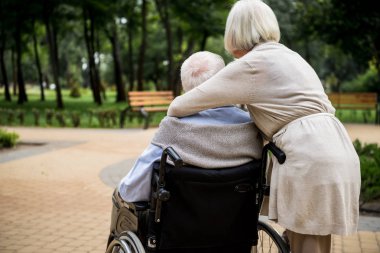 back view of senior woman with husband in wheelchair in park clipart