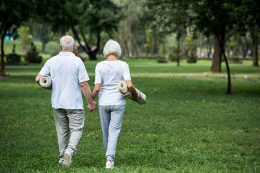 senior couple holding fitness mats while walking in park and holding hands clipart