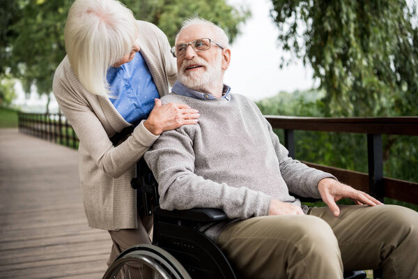 senior woman looking at smiling husband in wheelchair while holding hand on his shoulder