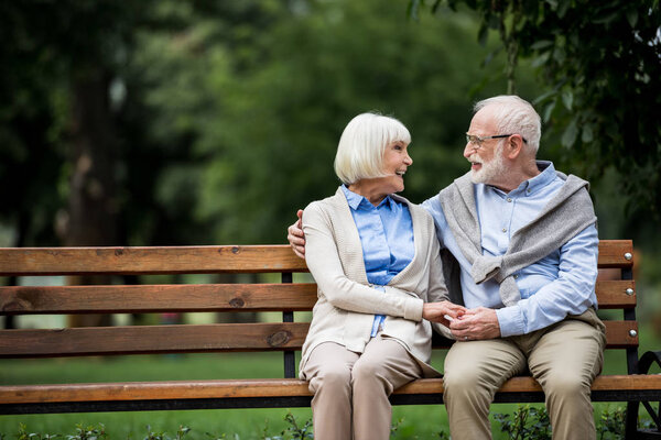 happy senior couple holding hands and looking at each other while sitting on wooden bench park