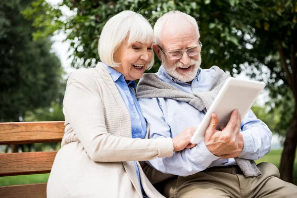 快乐的微笑的老夫妇使用数字平板电脑 而坐在公园的长凳上 — 图库照片