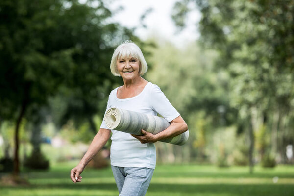 счастливая пожилая женщина с фитнес-ковриком, гуляющая в парке
