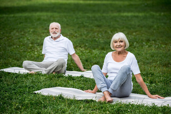 happy senior couple sitting in relaxation poses on yoga mats on green lawn 