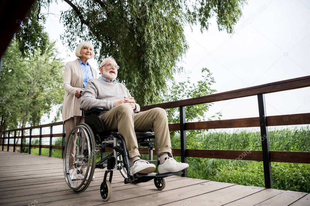 nice senior woman carrying husband in wheelchair while walking in park