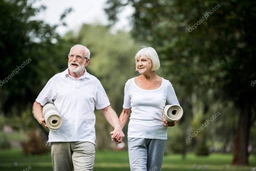 smiling senior couple with fitness mats walking in park and holding hands