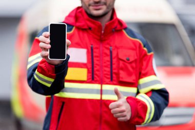 Partial view of paramedic holding smartphone with blank screen and showing thumb up clipart