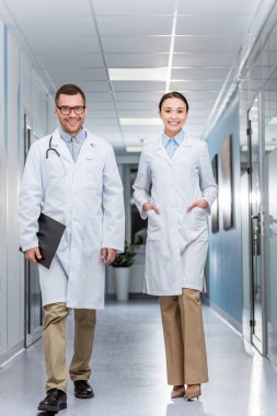 Happy doctors in white coats walking down hall together clipart
