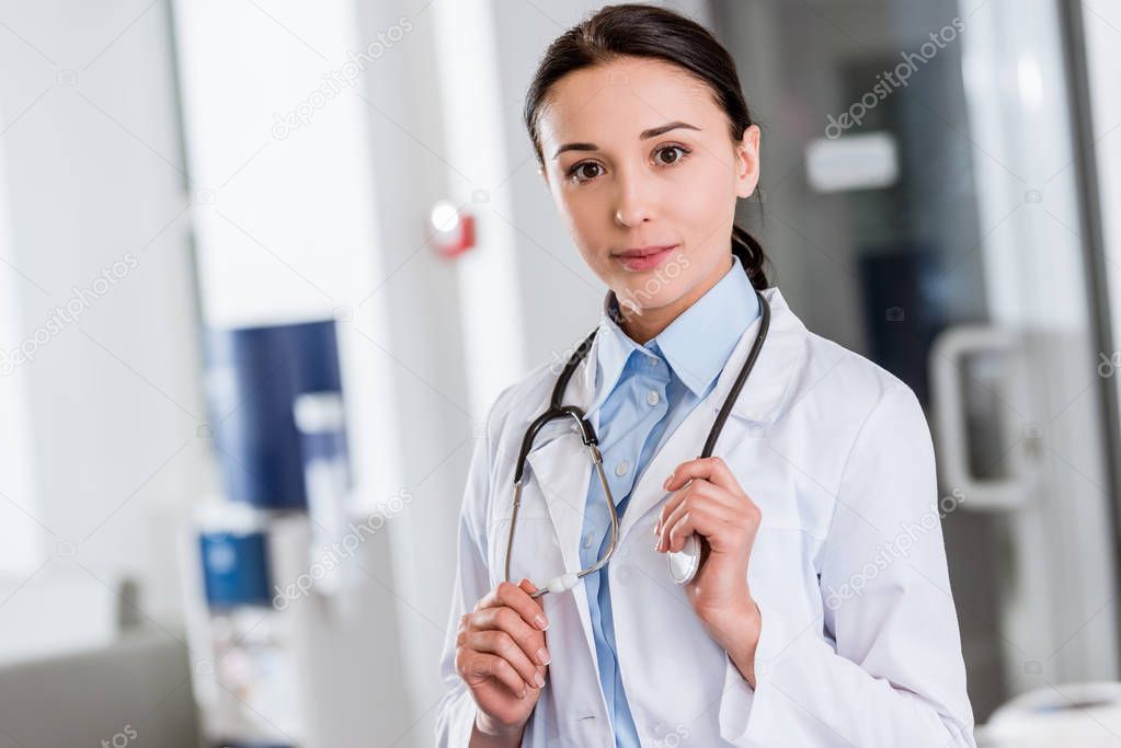 Young doctor in white coat touching stethoscope and looking at camera