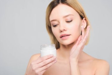 yüzünde kozmetik krem uygulamak ve plastik konteyner gri izole tutan güzel sarışın kadın