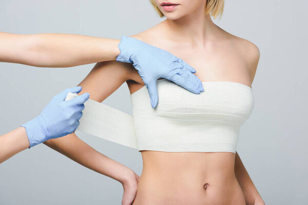 cropped view of plastic surgeon wrapping female breast with bandage, isolated on grey