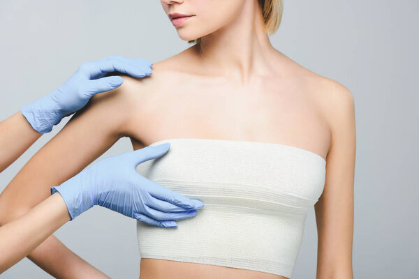 cropped view of plastic surgeon in latex gloves checking female breast in bandage, isolated on grey