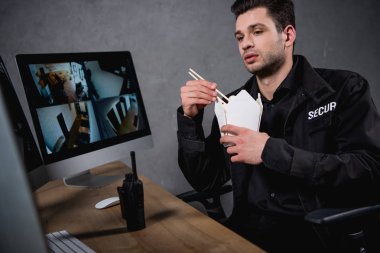 handsome guard in uniform eating takeaway food and looking at computer monitor  clipart