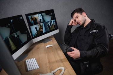 bored guard in uniform holding walkie-talkie and looking at computer monitor  clipart