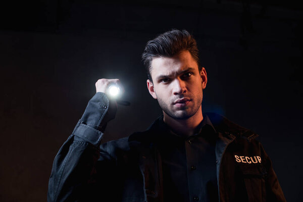 охранник в форме держит фонарик и смотрит в камеру
 