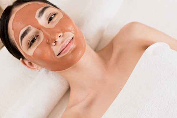 top view of beautiful woman lying with clay mask on face and looking at camera